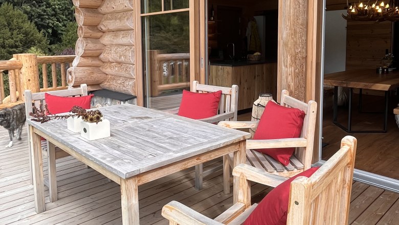 Holzterrasse mit Tisch und St&uuml;hlen, roten Kissen und einem Hund im Hintergrund.