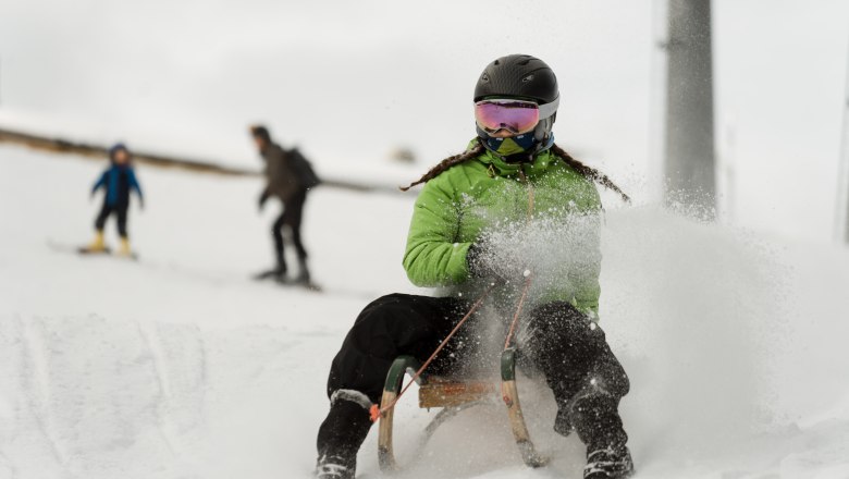 Person mit gr&uuml;nem Anorak und Helm f&auml;hrt auf einem Schlitten im Schnee.