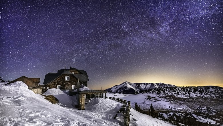 Sternenklare Nacht über einer verschneiten Berghütte und Berglandschaft.