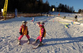 Zwei Kinder in Skiausrüstung auf einer Skipiste bei Sonnenschein.