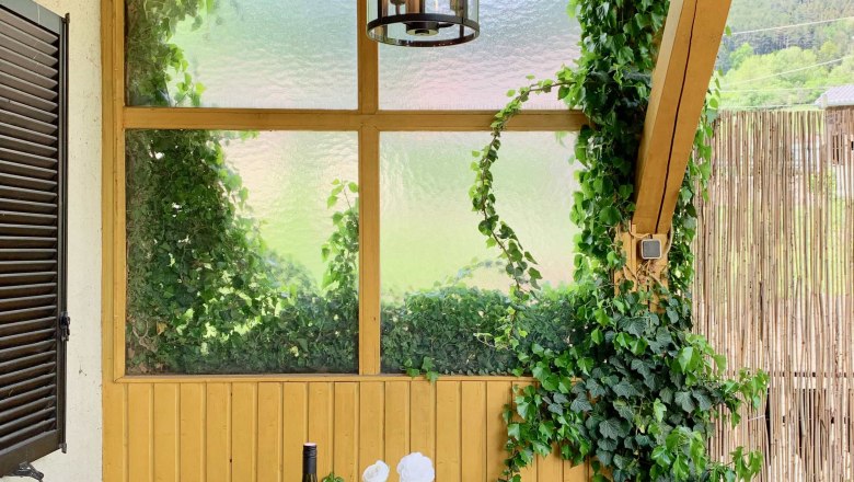 Ein gedeckter Tisch auf einer Terrasse mit Weinflasche, Geschirr und Kerzen, umgeben von Efeu und Holzwänden.