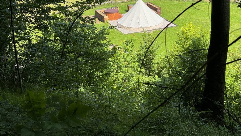 Blick auf ein Glamping-Zelt und einen Whirlpool auf einer Wiese, umgeben von B&auml;umen.