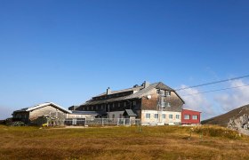 Panoramablick auf einen Bergh&uuml;tte mit umliegenden Gipfeln unter blauem Himmel.