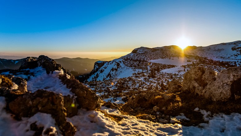 Sonnenuntergang über schneebedecktem Rax-Plateau mit Felsen im Vordergrund.