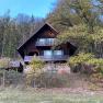Ein rustikales Holzhaus mit Balkon steht am Waldrand, umgeben von Bäumen und herbstlichem Laub.