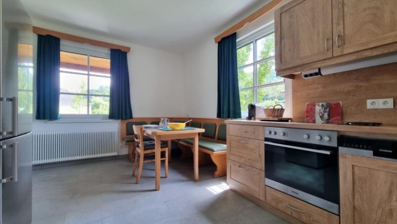 K&uuml;che mit Holzm&ouml;beln, Esstisch und Eckbank, zwei Fenstern mit gr&uuml;nen Vorh&auml;ngen.