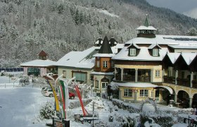 Ein verschneites Hotel im alpinen Stil mit Türmen und Balkonen, umgeben von schneebedeckten Bäumen und Hügeln.