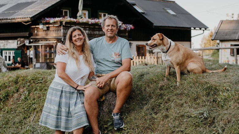 Ein Paar sitzt auf einem Hügel vor einer Hütte mit einem Hund daneben.