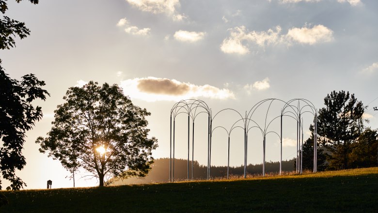 Ein Baum und eine Metallskulptur auf einer Wiese bei Sonnenuntergang.