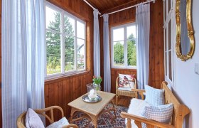 Ein gemütlicher Wintergarten mit Holzwänden, Korbmöbeln, blauen Vorhängen und einem Tisch mit Kerze und Blumen.