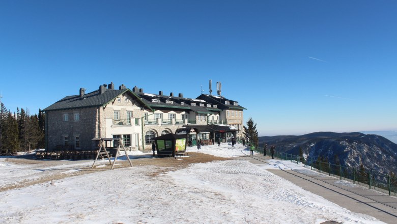 Raxalm Berggasthof im Winter mit schneebedecktem Boden und blauem Himmel.