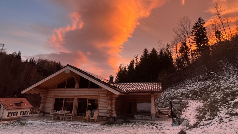 Ein Holzchalet im Winter mit schneebedecktem Boden und einem dramatischen, orangefarbenen Himmel im Hintergrund.