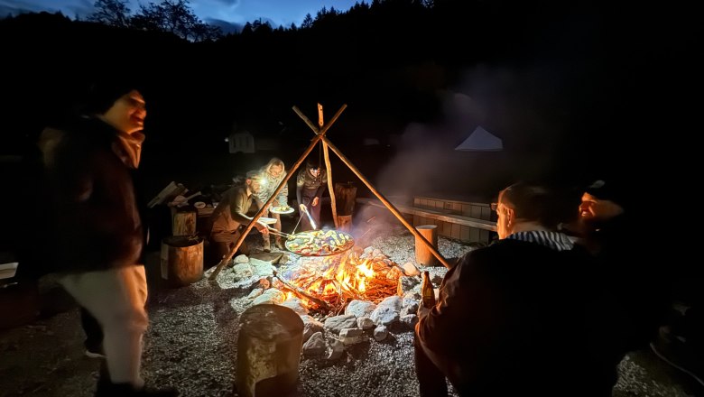 Menschen sitzen nachts um ein Lagerfeuer im Freien, umgeben von Bäumen.