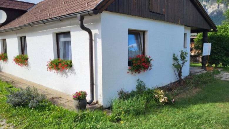 Weißes Haus mit roten Blumen in Blumenkästen vor den Fenstern, grüne Wiese im Vordergrund.