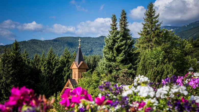 Kirchturm inmitten von B&auml;umen und Blumen vor einer Berglandschaft.