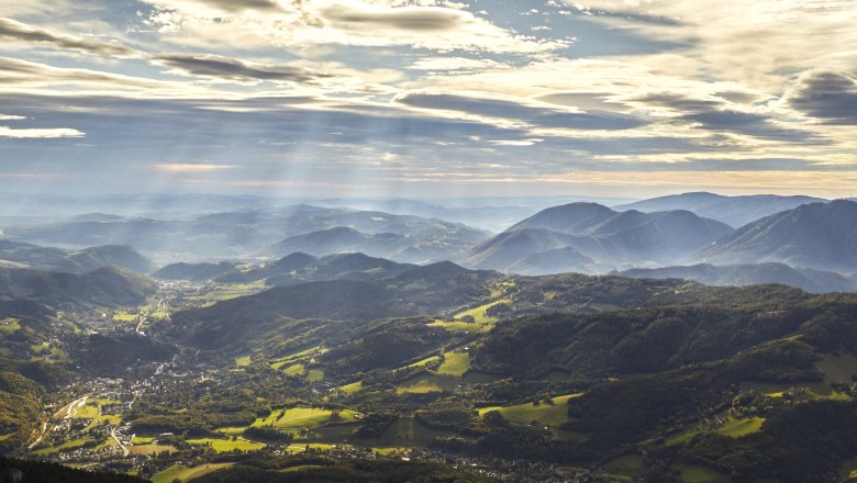 Panoramablick auf die Rax-Alpen mit bewaldeten Hügeln und Sonnenstrahlen durch Wolken.