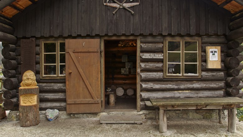 Eine rustikale Holzh&uuml;tte mit offener T&uuml;r, Holzschnitzerei und Tisch davor.