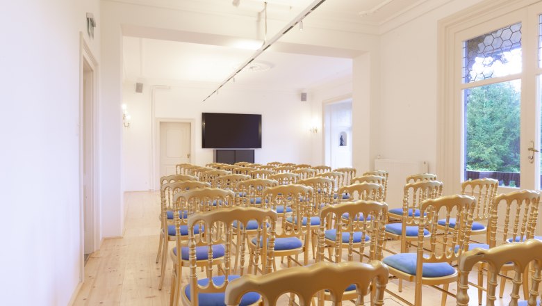 Ein heller Salon mit Stuhlreihen und einem gro&szlig;en Bildschirm an der Wand.