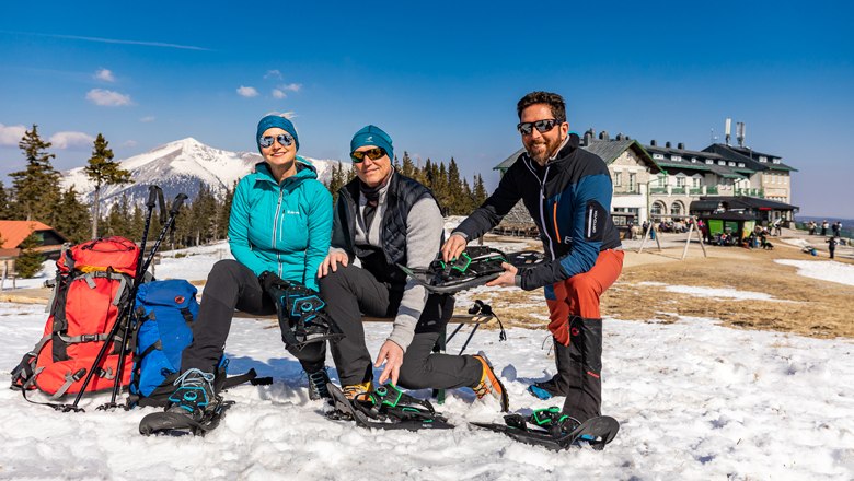 Drei Personen sitzen im Schnee mit Schneeschuhen vor einem Berg und einem Gebäude.