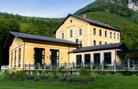 Außenansicht eines gelben Gebäudes mit Garten und Wald im Hintergrund.