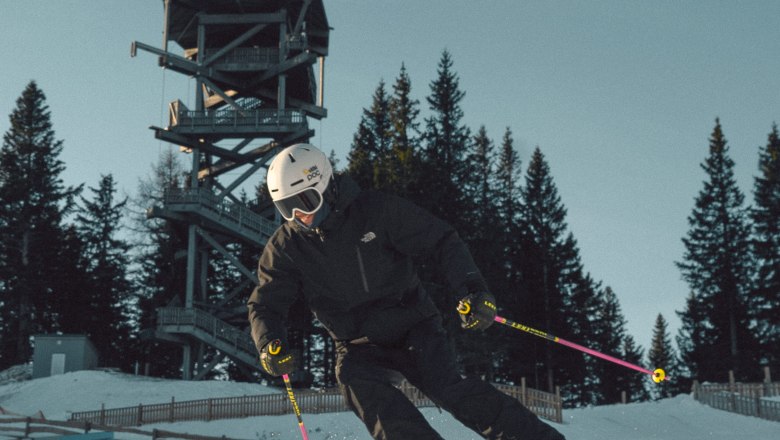 Skifahrer in schwarzer Kleidung fährt auf präparierter Piste am Semmering Hirschenkogel, im Hintergrund ein Aussichtsturm und Bäume.