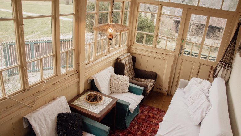 Gemütlicher Wintergarten mit Sesseln, Sofa und Tisch, umgeben von großen Fenstern mit Blick ins Freie.