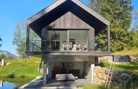 Modernes Haus mit Holzfassade und Balkon, umgeben von Natur und einem kleinen Teich.