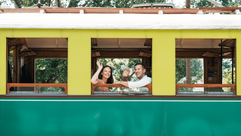 Zwei Personen winken aus einem gelb-grünen Zugfenster.