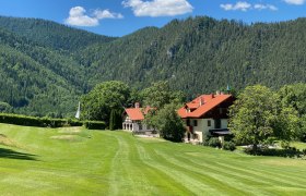 Ein Clubhaus mit rotem Dach in einer gr&uuml;nen, bergigen Landschaft unter blauem Himmel.