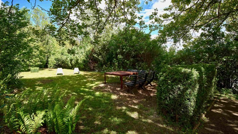 Ein grüner Garten mit Bäumen, Sträuchern, zwei weißen Liegestühlen und einer Sitzgruppe mit Tisch im Schatten.