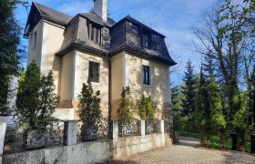Eine zweistöckige Villa mit Mansardendach, umgeben von Bäumen und einem schmiedeeisernen Zaun.