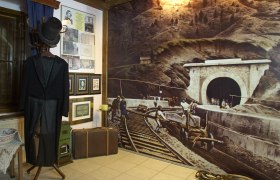 Innenansicht des Ghega Museums mit historischem Anzug, Bildern und einer großen Wandmalerei eines Eisenbahntunnels.