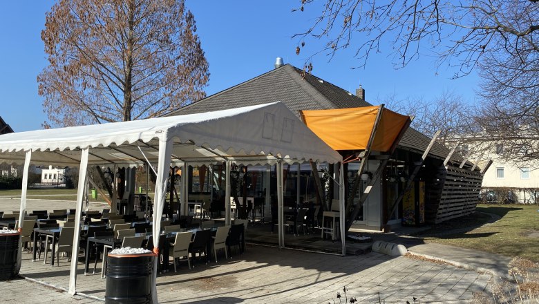 Ein Caf&eacute; mit Au&szlig;ensitzbereich im Eduard-Garderer-Park, umgeben von kahlen B&auml;umen und blauem Himmel.